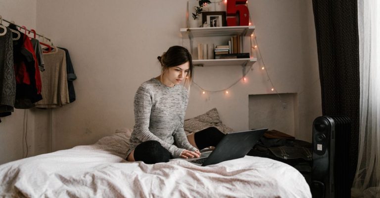 A woman in a gray sweater working on a laptop in a cozy bedroom. Ideal for remote work or study themes.