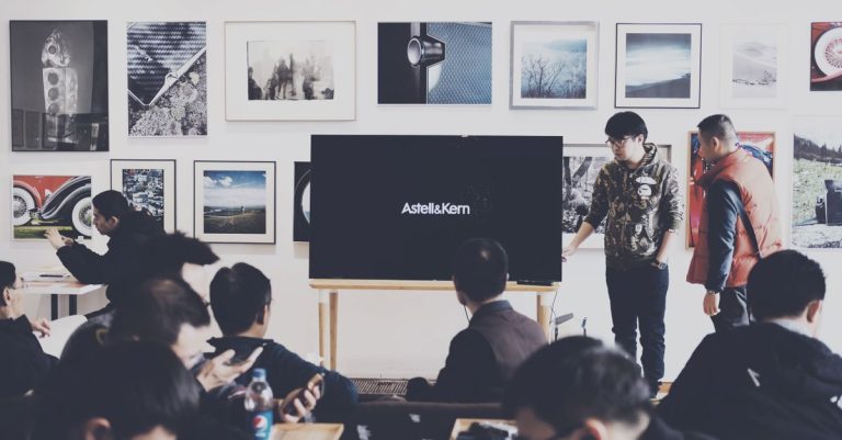A group of adults attending a casual business presentation in a modern art space, featuring a TV display and framed art.
