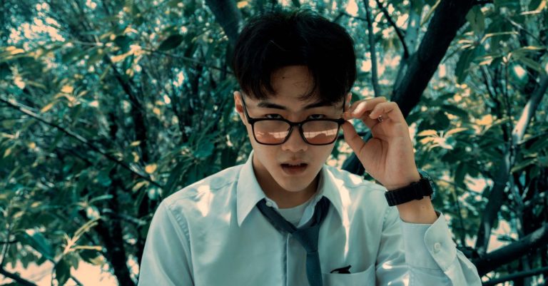 Portrait of a young man with glasses in a white shirt and tie outdoors among green trees.