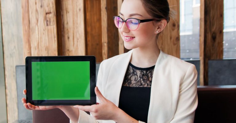 Smiling woman in corporate attire holding a tablet with a green screen indoors.