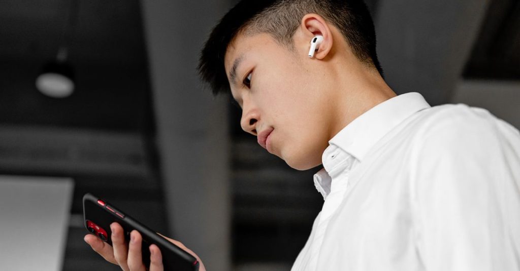 Side view of a young man wearing wireless earbuds and using a smartphone indoors.