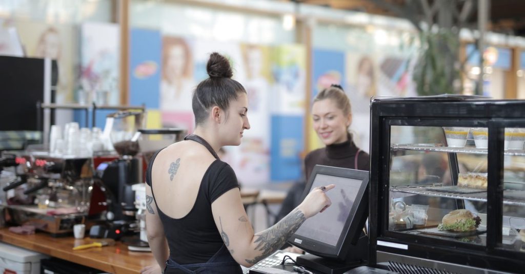 Female cashier with tattoos helps a customer with a transaction at a cafe counter.